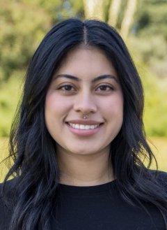 Woman with dark hair parted in the middle, wearing a black shirt, standing in front of greenery.